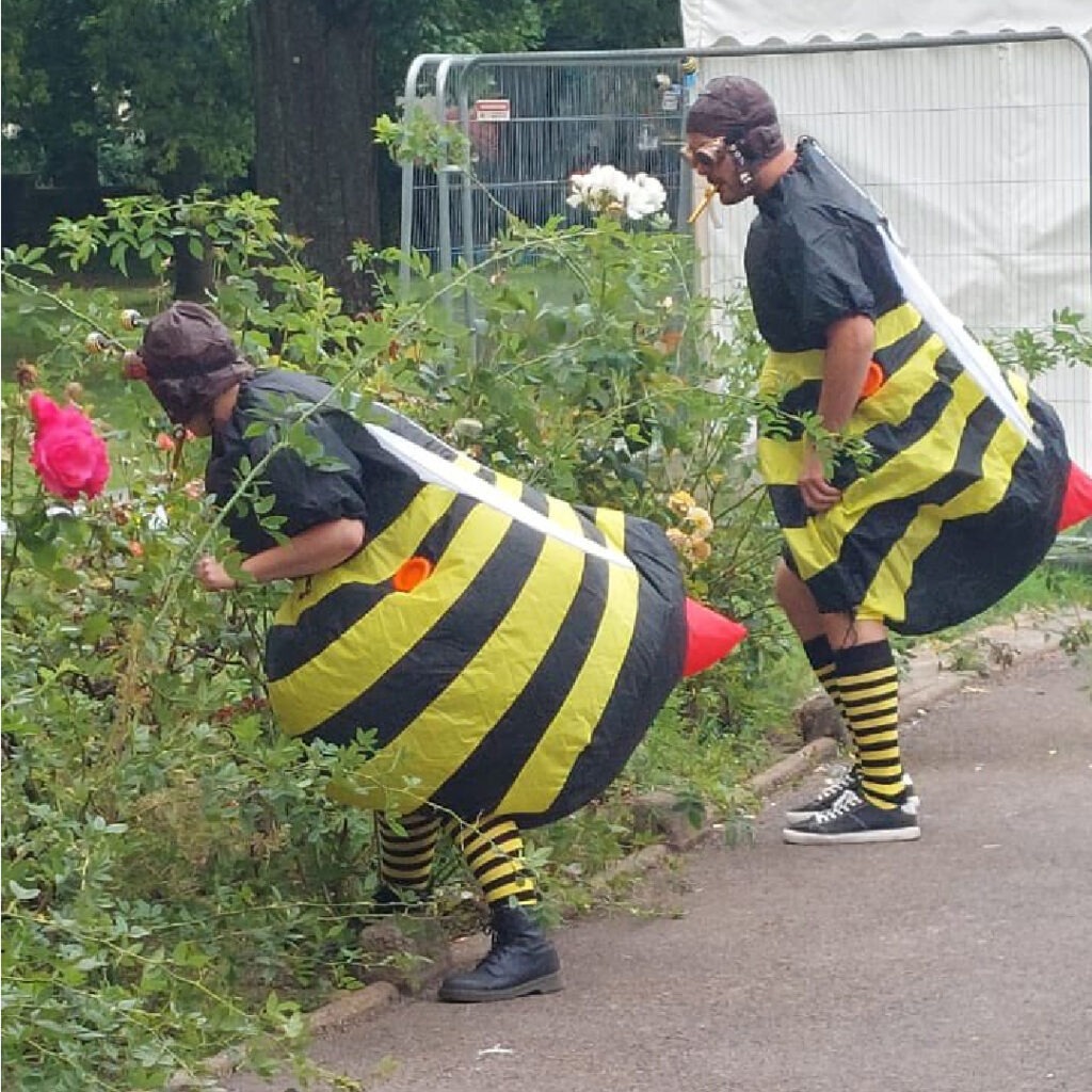 Two performers dressed as bees smell some pink rose bushes